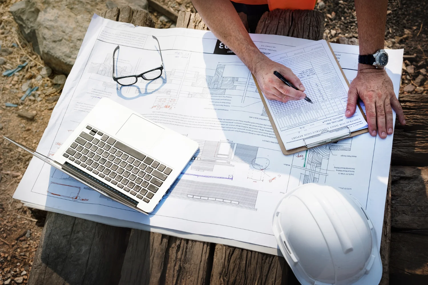 Construction worker reviewing architectural blueprints with a laptop, safety helmet, and clipboard at an outdoor worksite.